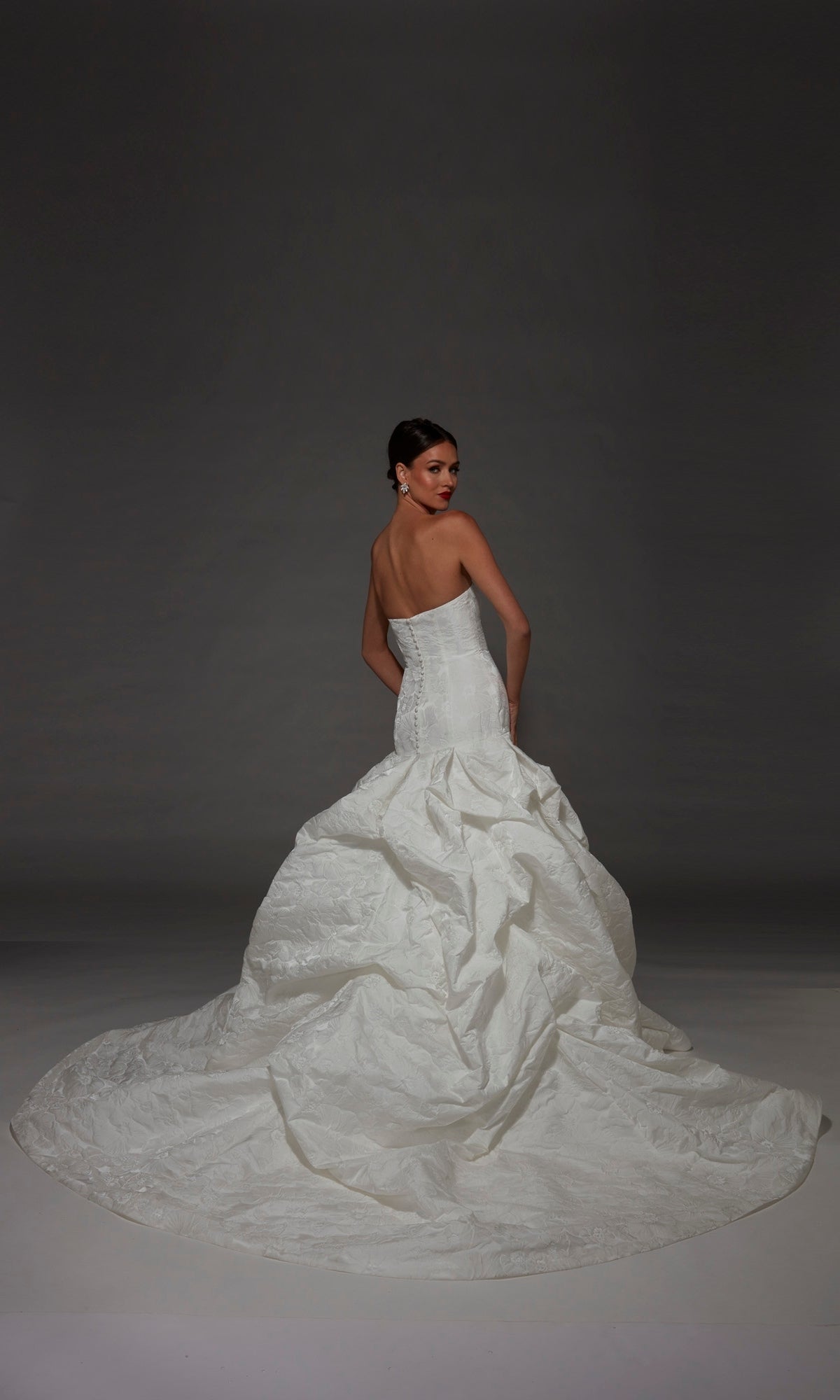 Wearing the Long Wedding Dress: Alyce 7185, a woman poses with her back turned, highlighting the gownโs strapless jacquard design and sweeping textured train against a dark background.